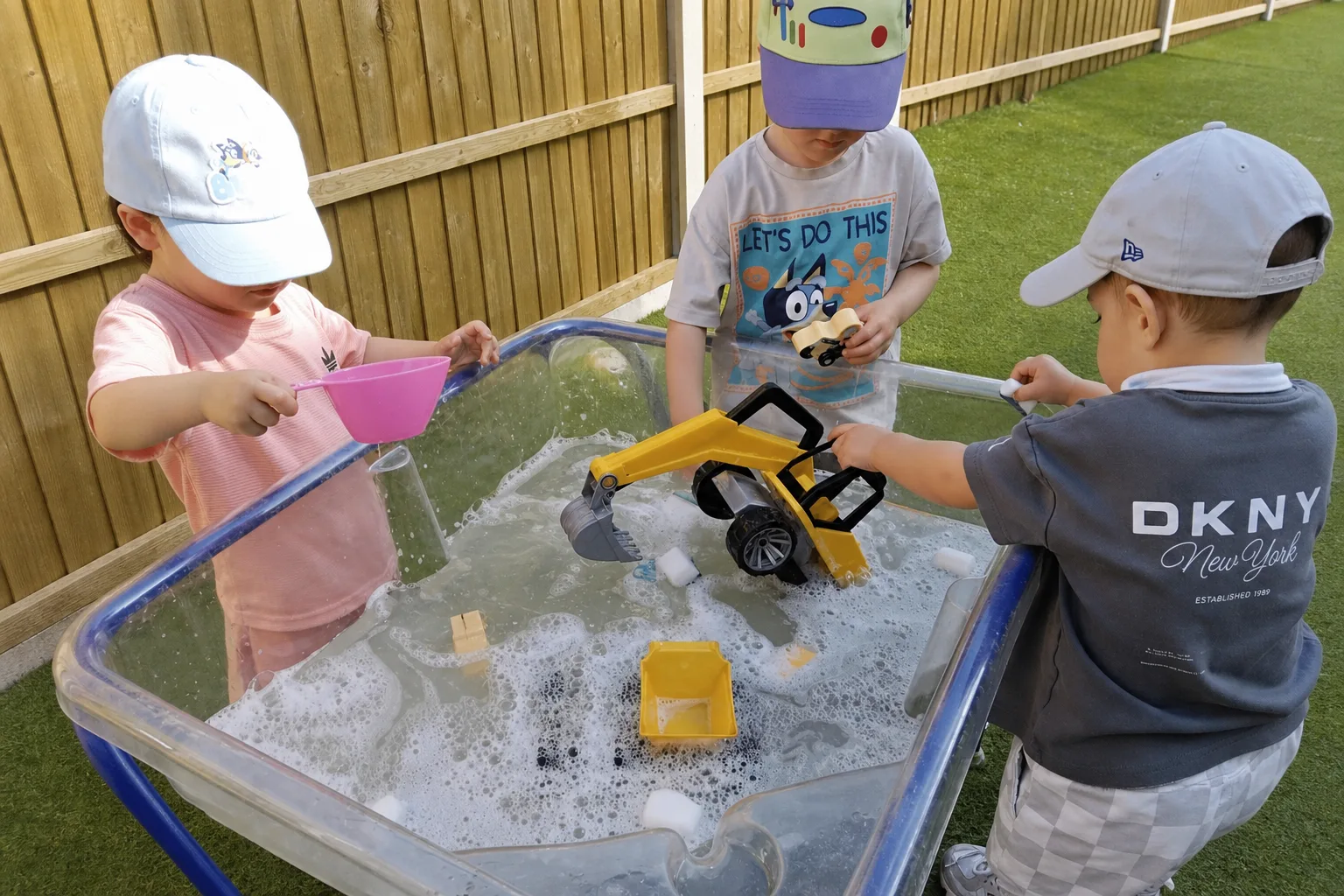 Three young children playing together with vehicles, cups and bubbly water at an outdoor water table
