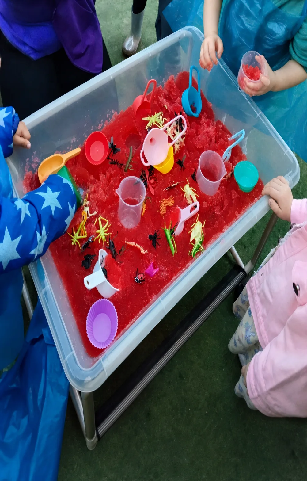 A red water sensory tray with toy minibeasts, cups and scooping tools